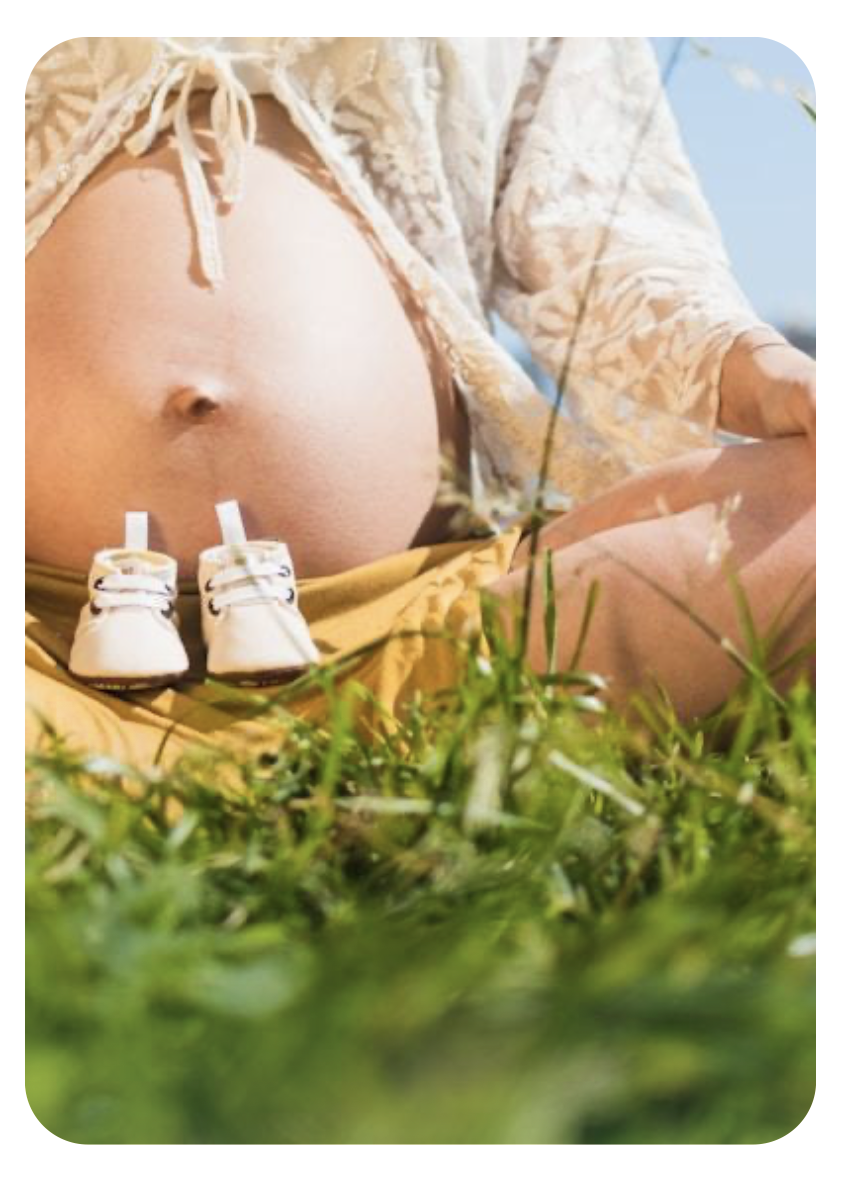 Pregnant woman sitting on grass with baby shoes